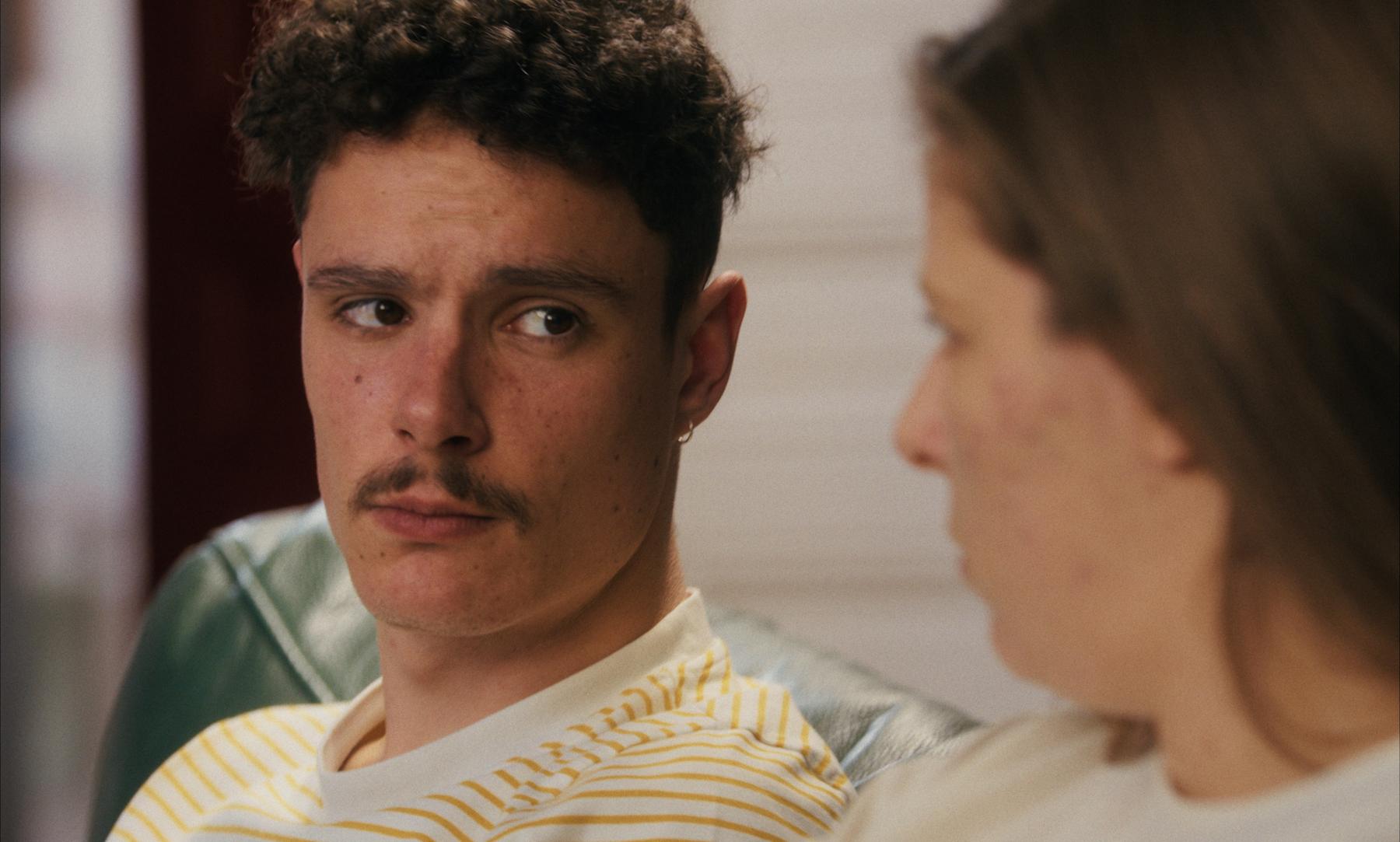 Photogramme : regard en coin d'un jeune homme à moustache sur le canapé.