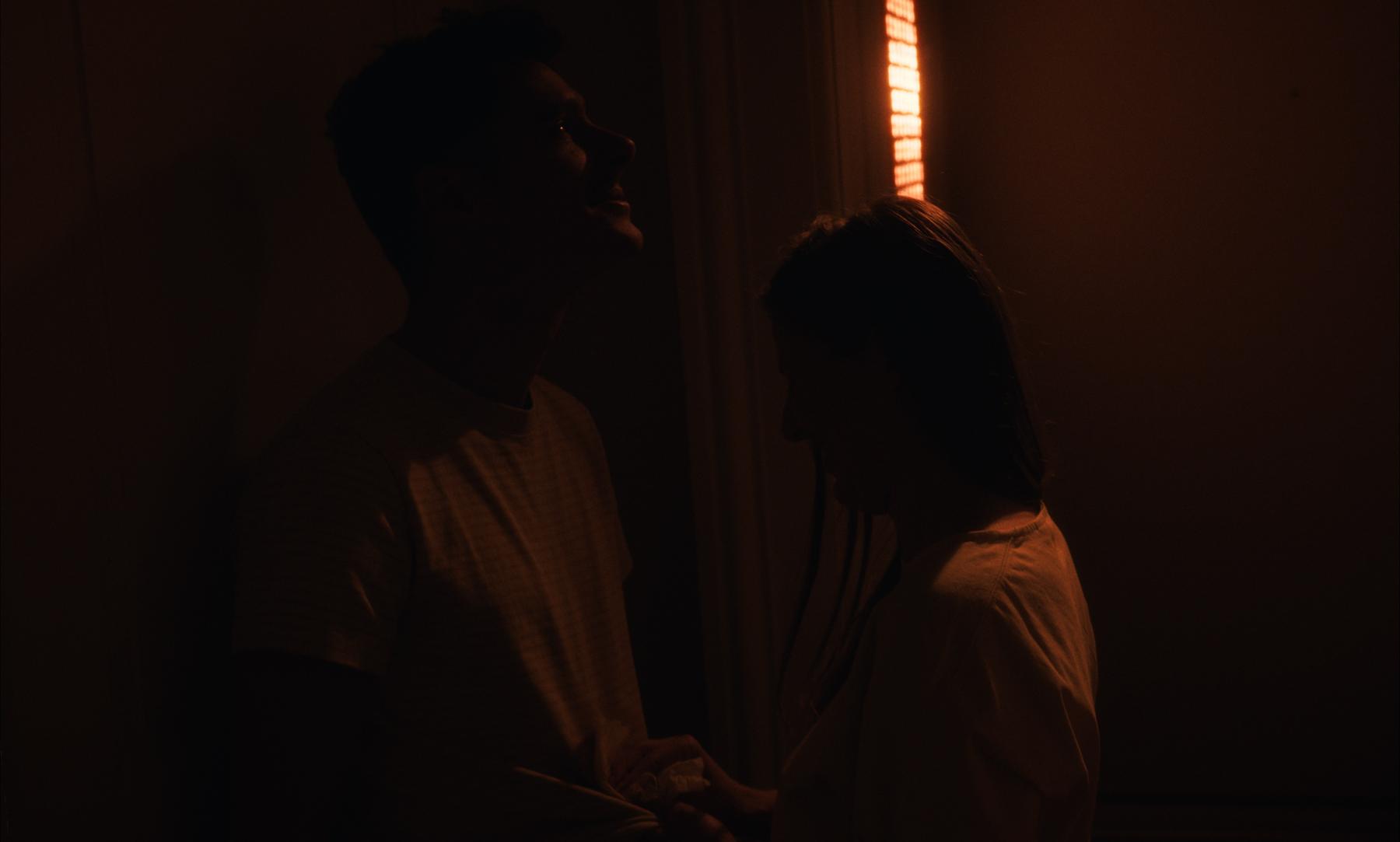 Photogramme : un couple dans la pénombre, un rai de lumière orange entre eux.