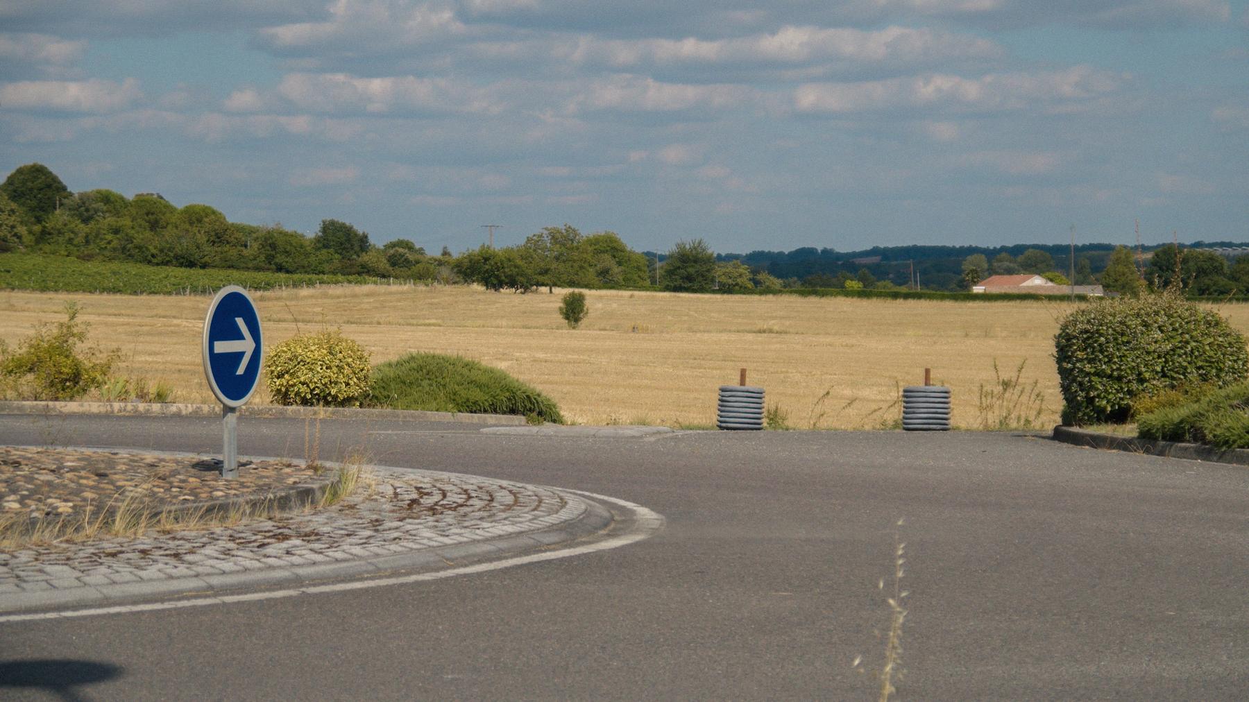 Repérage : un rond-point désert au milieu des champs moissonnés, une flèche bleue plantée au centre.