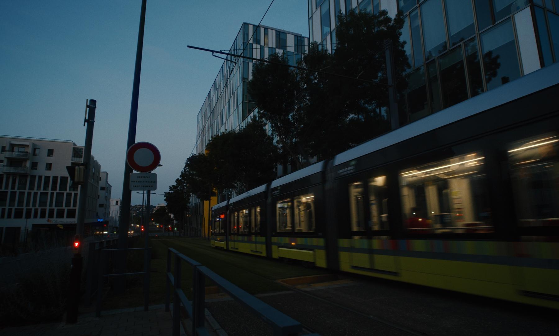 Photogramme : le tramway jaune passe de nuit devant les façades de verre.